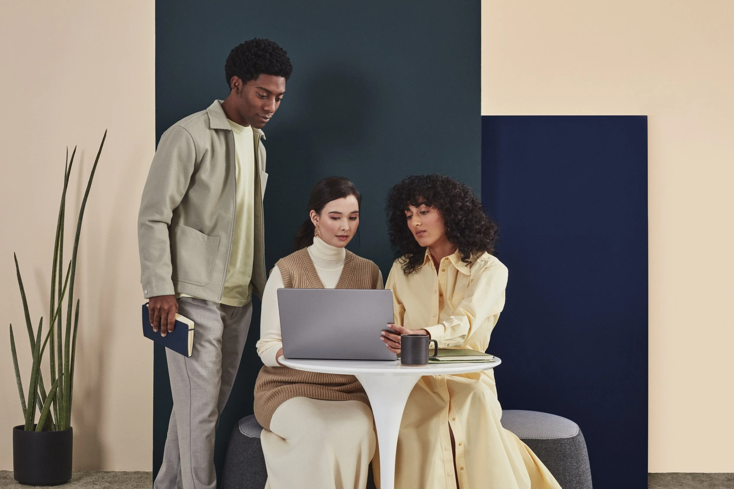 Three young women working together around a laptop in a modern office setting. One woman is sitting with a notebook and a coffee mug, another woman is sitting and using the laptop, and a third woman is standing and holding a notebook. The background 