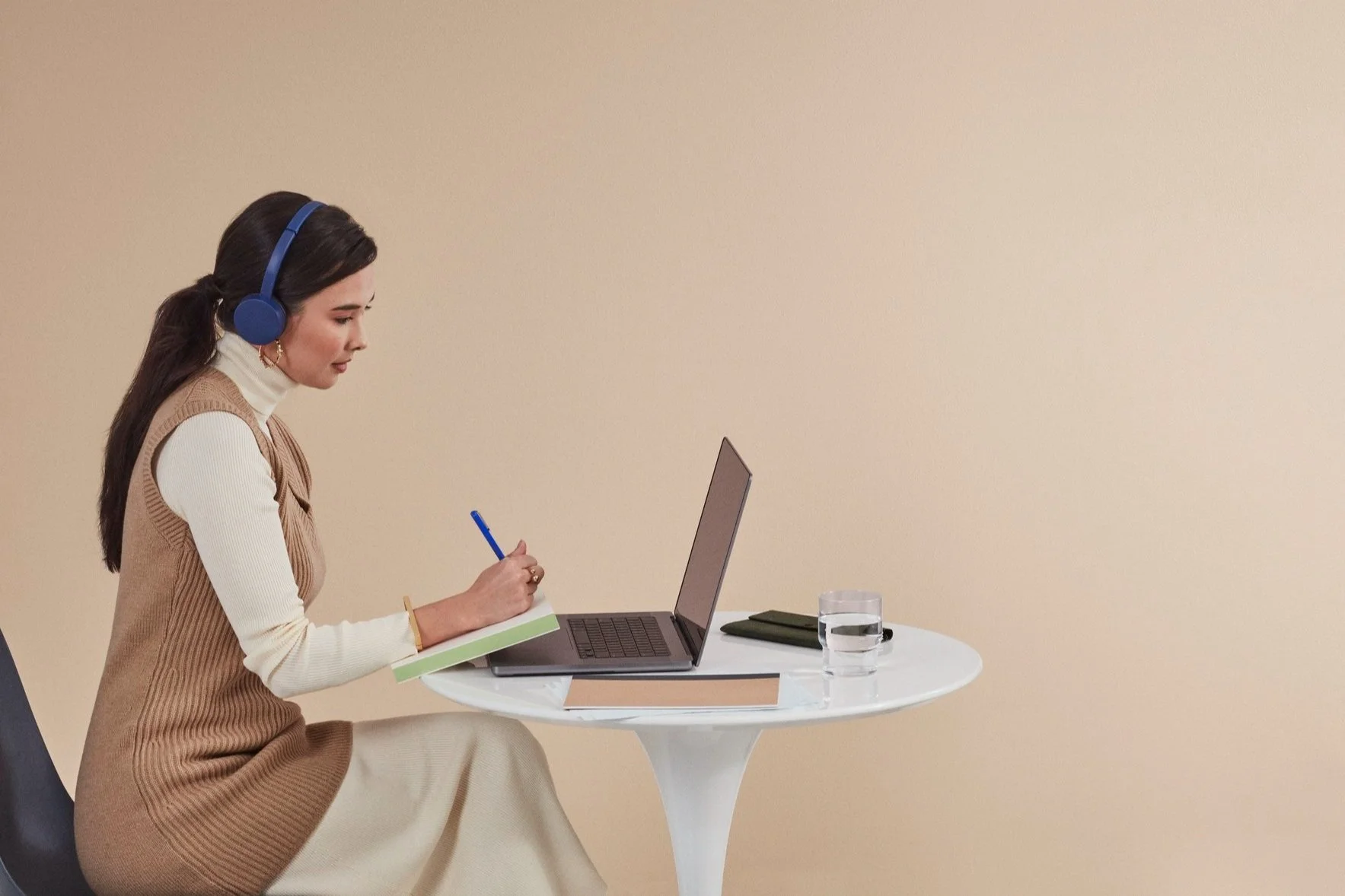 A woman sitting at a white round table with a laptop, notebook, a glass of water, a pen, and a wallet. She is wearing a beige vest, white turtleneck, and blue headphones, and is writing in a notebook.