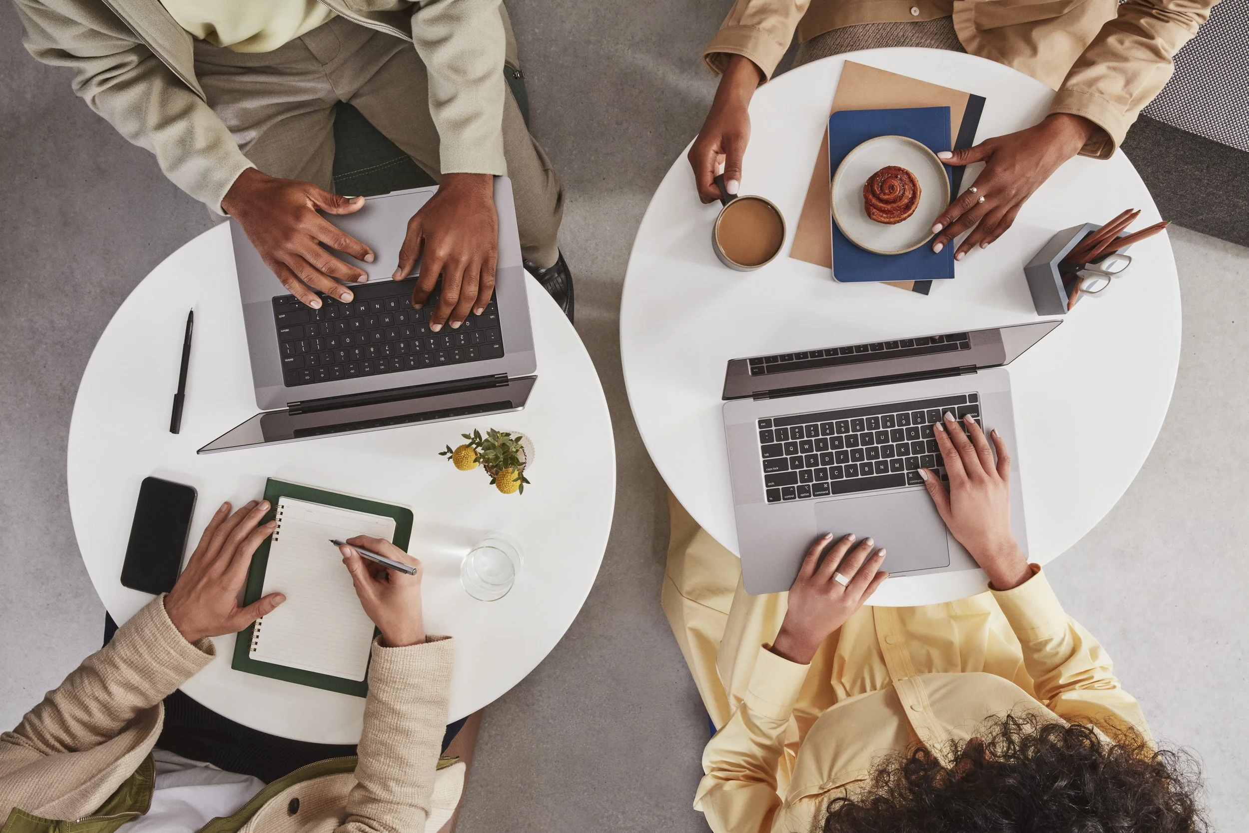People working at a white round table with laptops, notebooks, coffee, and snacks, seen from above.