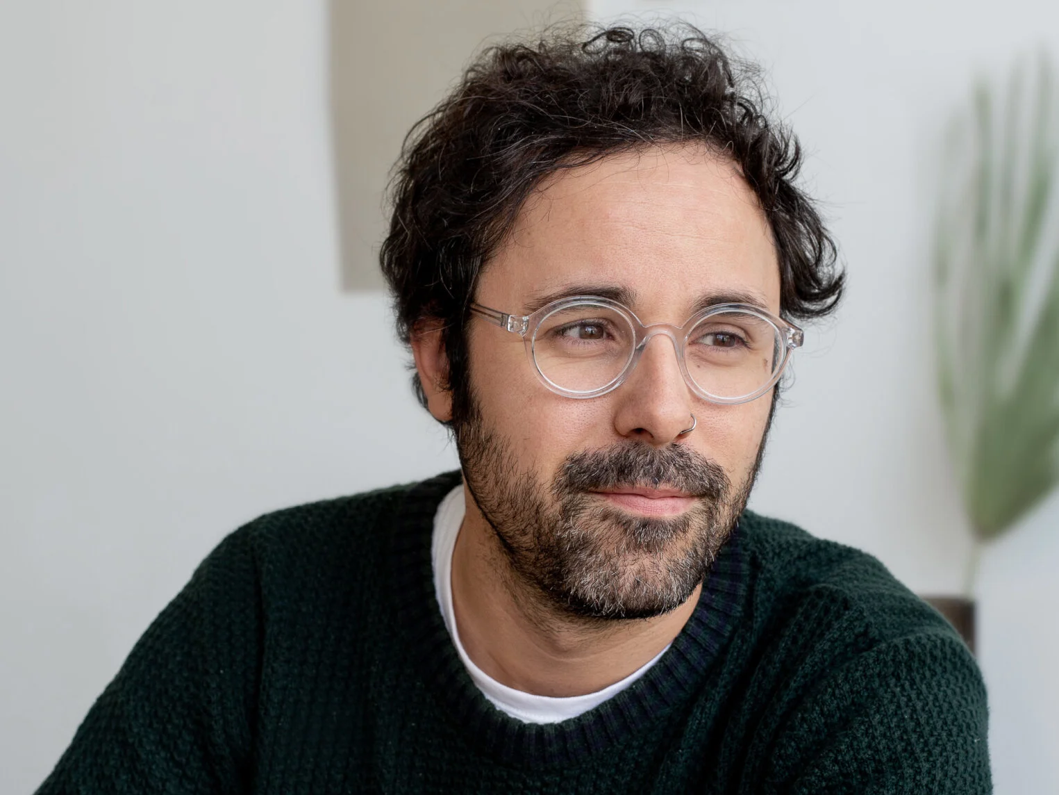 A man with curly dark hair, glasses, and a beard, wearing a black sweater, sitting indoors with a plant in the background.