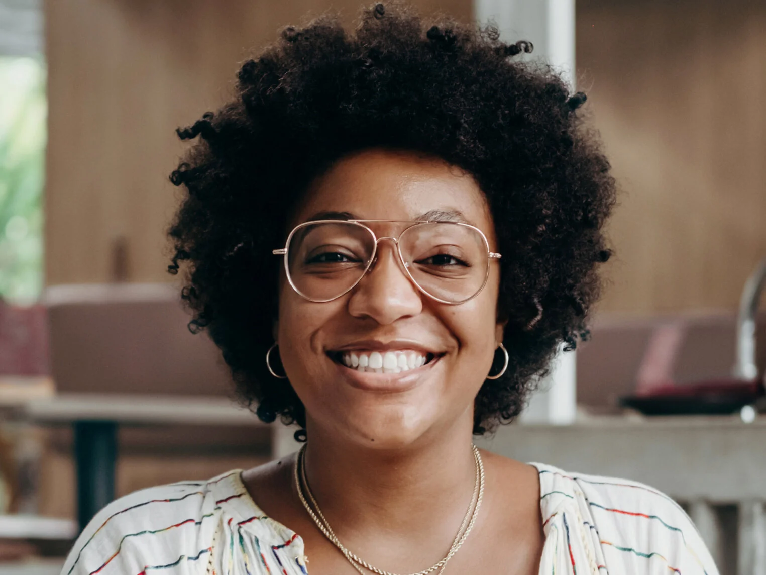 Close-up of a smiling African American woman with curly hair, wearing glasses, hoop earrings, and layered necklaces, in a bright indoor setting.