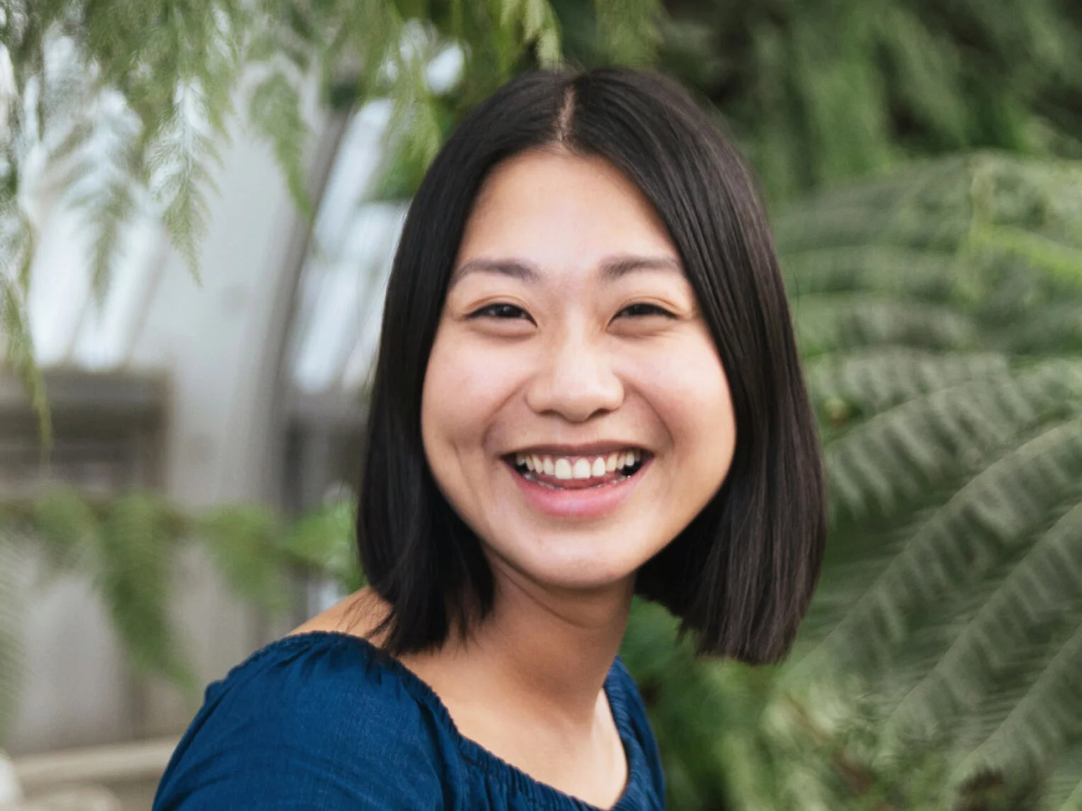A young woman with short black hair smiling in a greenhouse or indoor garden with green plants in the background.
