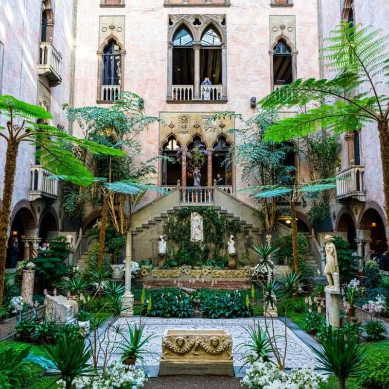Interior courtyard of a historic building with a lush garden, statues, and ornate architecture. Features arched windows, balconies, and greenery.