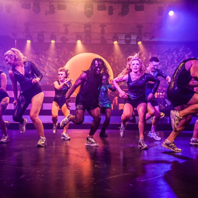 The company of Wake dancing on a purple-lit stage